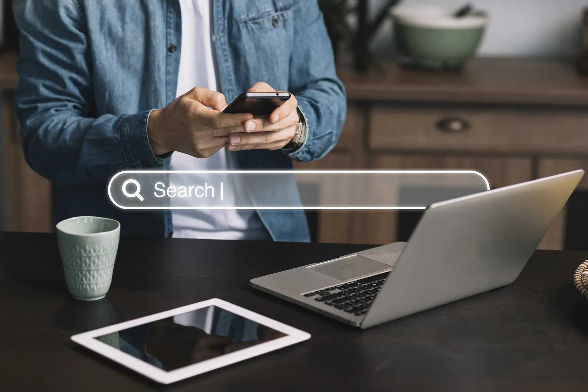 Personne tapant une recherche sur son smartphone, barre de recherche en surimpression, ordinateur portable et tablette sur le bureau